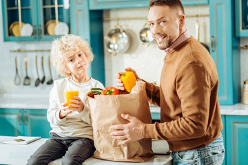 Fresh juice. Nice pleasant young boy sitting on the table and holding juice while being with his father in the kitchen