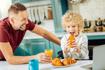 Tasty croissant. Happy positive joyful boy holding a croissant and taking a bite while standing with his father in the kitchen