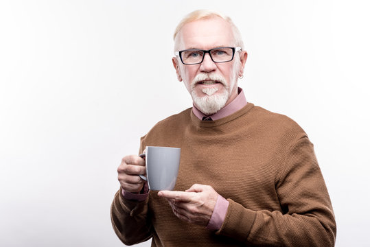 Delicious Coffee. Handsome Elderly Man In Eyeglasses Holding A Cup Of Coffee In One Hand And Supporting It With The Other While Posing Isolated On A White Background