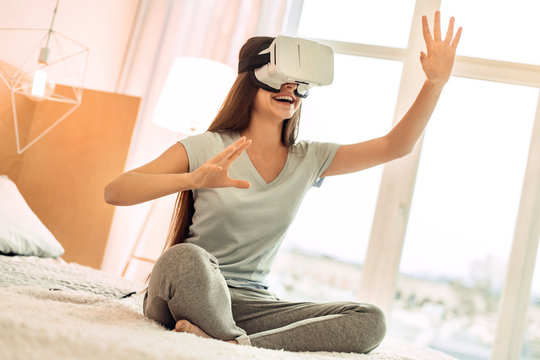 Best game. Joyful teenage girl sitting on her bed and playing with a virtual reality headset while smiling brightly