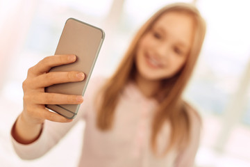 Helpful gadget. The focus being on the phone in the hands of a pretty fair-haired teenage girl standing in the room and taking a selfie while smiling