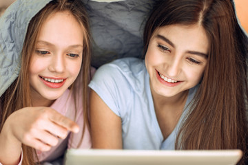 Movie night. Pleasant teenage girl lying on the bed, being covered with a blanket, and watching a film on the tablet while one of the girls pointing at it with her finger