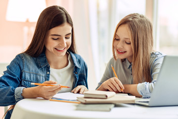 Pleasant cooperation. Upbeat teenage girls sitting at the table and doing their home assignment, helping each other with it while smiling