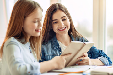 Knowledge is power. Charming teenage girls sitting at the table in the living room and reading a book together while doing their home assignment