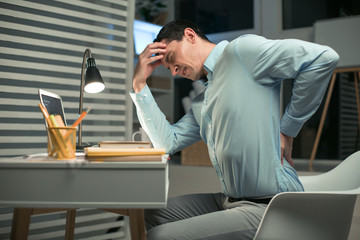 Terrible pain. Young tired male employee sitting in the chair while closing eyes and touching his head and back