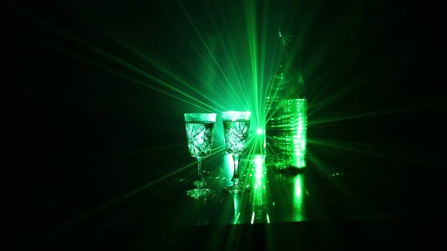 Two glasses of Vodka with bottle on dark foggy club style background with glowing lights (Laser, Stobe) Multi colored. Club drinks theme decoration. Empty space. Selective focus