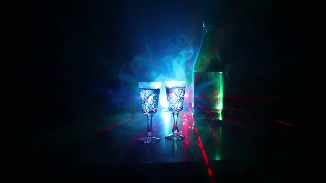 Two glasses of Vodka with bottle on dark foggy club style background with glowing lights (Laser, Stobe) Multi colored. Club drinks theme decoration. Empty space. Selective focus
