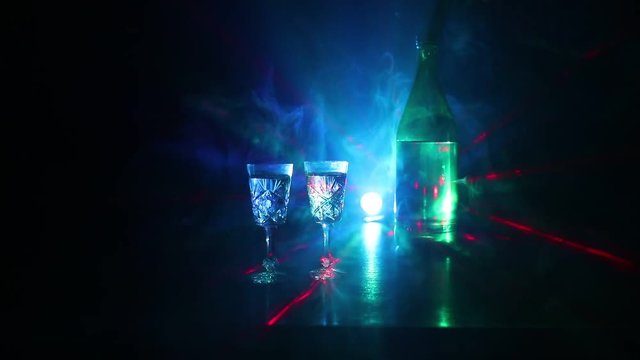 Two glasses of Vodka with bottle on dark foggy club style background with glowing lights (Laser, Stobe) Multi colored. Club drinks theme decoration. Empty space. Selective focus