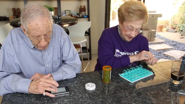 Husband reads smartphone while wife fills pill box with medication