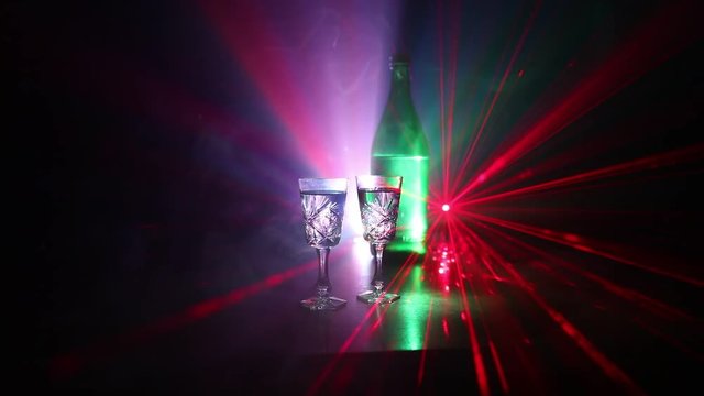 Two glasses of Vodka with bottle on dark foggy club style background with glowing lights (Laser, Stobe) Multi colored. Club drinks theme decoration. Empty space. Selective focus