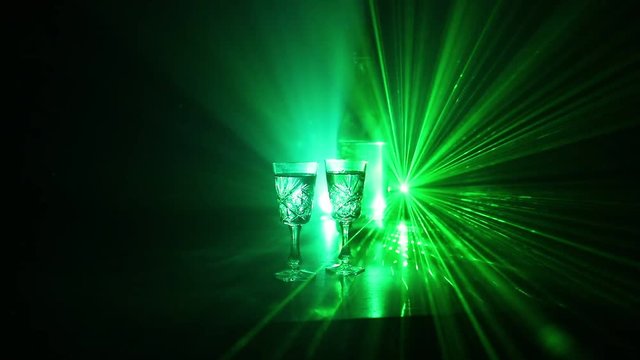 Two glasses of Vodka with bottle on dark foggy club style background with glowing lights (Laser, Stobe) Multi colored. Club drinks theme decoration. Empty space. Selective focus