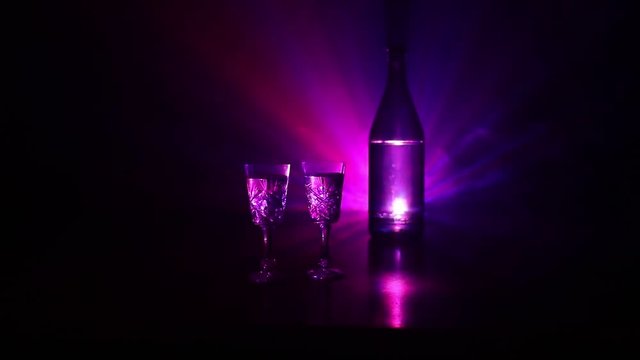 Two glasses of Vodka with bottle on dark foggy club style background with glowing lights (Laser, Stobe) Multi colored. Club drinks theme decoration. Empty space. Selective focus