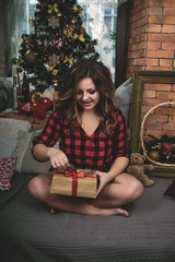young joyful beautiful girl in a red shirt opens her gift on the background of a Christmas tree, windows and walls of red bricks