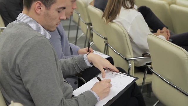 The two businessmen sit among their colleagues on business presentation and take notes on papers. The one man wears gray jumper and writes by the pen and the other male is in blue suit. Other business