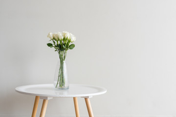 White roses in glass vase on small white table against beige wall