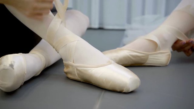 Two ballerinas are tying tape of pointe shoes sitting on floor in school. Graceful dancers tie ribbon of shoes before practice in modern ballet studio. Young ladies in silk outfit sit on parquet and