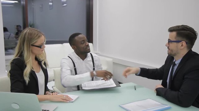 Multi-ethnic businesspeople have just signed the contract in the office. Businessman in glasses is giving the notepad with paper documents to the blond female manager and shaking hand with african