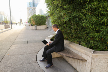 Businessman typing by laptop outside near green plant in   and looking at watch. Concept of ecology...