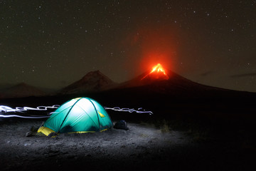 Illuminated tourist camp on background eruption volcano, light from headlights around tent