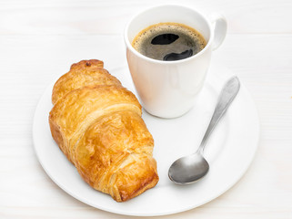 Cup of coffee, croissant and teaspoon on the white plate.