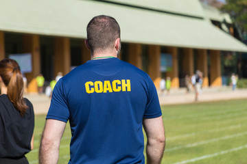 Back side of a male athlete coach in deep blue t-shirt with yellow COACH word written on its back