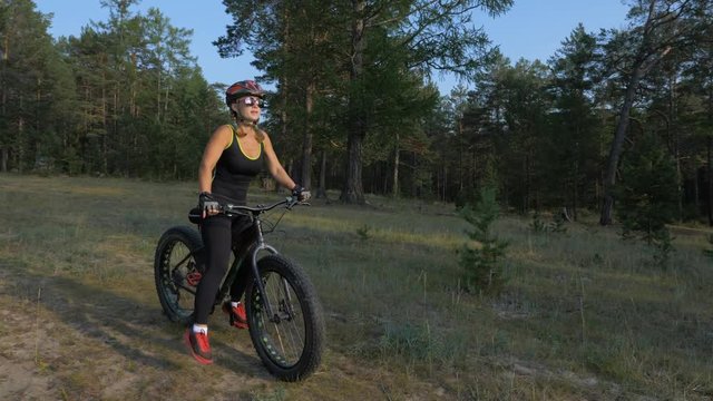 Fat bike also called fatbike or fat-tire bike in summer riding in the forest. Beautiful girl and her bicycle in the forest. She enters the frame and takes off her helmet and glasses.