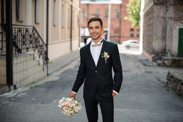 groom in a black suit walking down the street holding a pink bouquet