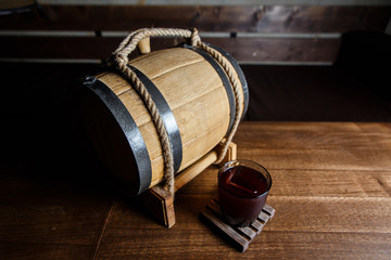 An underexposed horizontal image of a red alcoholic drink in a cocktail glass, on a wooden glass-holder on a table, a wooden barrel near by. Selective focus.