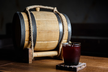 An underexposed horizontal image of a red alcoholic drink in a cocktail glass, on a wooden glass-holder on a table, a wooden barrel near by. Selective focus.