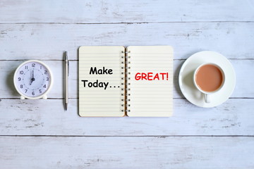 Top view of notebook written with 'MAKE TODAY GREAT!' with table clock pen and a cup of coffee on white wooden background.