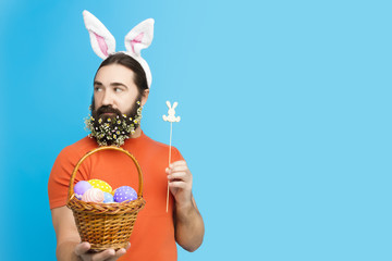 Happy Easter concept. Nice kind muscle man male with spring's flower's beard, white ears of rabbit and basket with colorful easter eggs and wooden rabbit in green t-shirt isolated on blue background