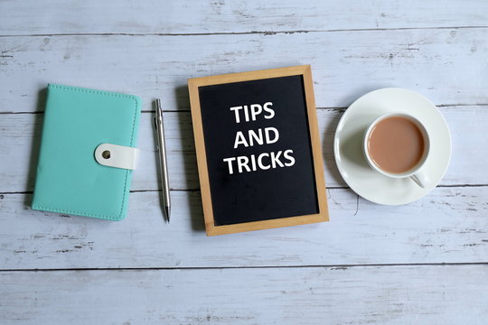 Top view of notebook,pen,a cup of coffee, and blackboard written with 'TIPS AND TRICKS' on white wooden background.