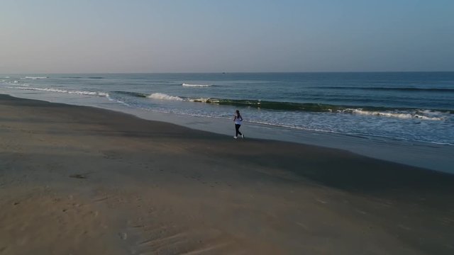 A young girl runs along a sandy beach in the morning next to the beautiful surf waves. Athletic young woman runs on the beach along the sea with long flowing hair. Aerial view.