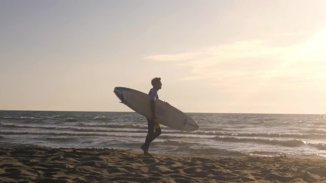 Young man surfer in wetsuit picks up surf board at the beach walking on the sand along sea shore looking at the ocean at sunset gimbal dolly steadycam