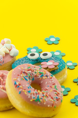 Many multicolored funny glazed donuts surrounded by artificial flowers on yellow background. Vivid hapyp contrast.