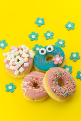 Many multicolored funny glazed donuts surrounded by artificial flowers on yellow background. Vivid hapyp contrast.