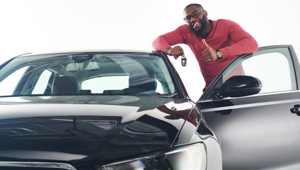 Happy handsome african man showing car keys in near his newly bought vehicle car smiling cheerfully copyspace owner ownership sales driving consumerism private taxi concept studio shot