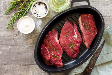 Fresh meat. Raw steak beef on a cast-iron grill frying pan, olive oil, spices and fresh rosemary on the kitchen table. Flat lay. Top view.
