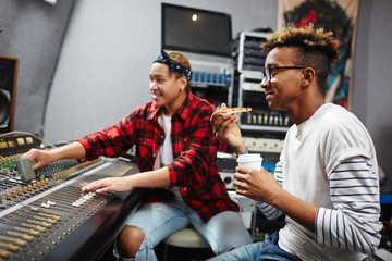 Young sound operator with drink and slice of pizza having lunch while his colleague working by soundboard