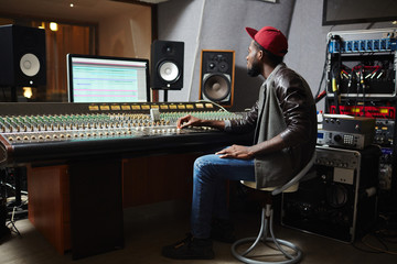 Young hipster sitting on chair in front of monitor and soundboard in studio of records