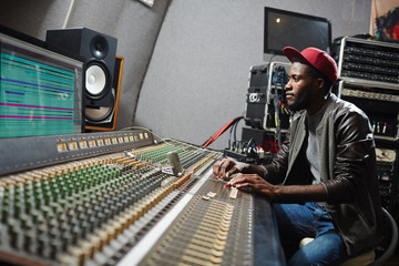 Young professional sitting by soundboard and mixing sounds while adjusting records