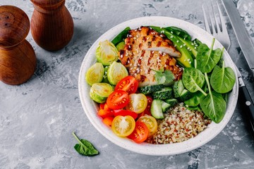 Healthy buddha bowl lunch with grilled chicken, quinoa, spinach, avocado, brussels sprouts, tomatoes, cucumbers  on dark grey background.