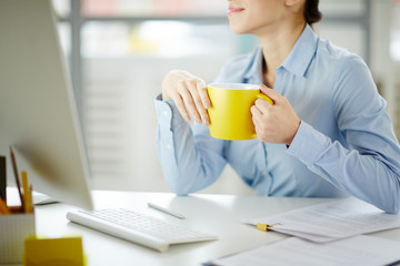 Young woman with yellow mug sitting by desk in office in front of computer monitor