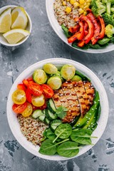 Healthy buddha bowl lunch with grilled chicken, quinoa, spinach, avocado, brussels sprouts, tomatoes, cucumbers  on dark grey background.