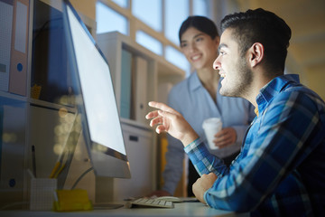 One of creative designers sitting in front of computer monitor and showing curious design project to colleague while discussing it with her