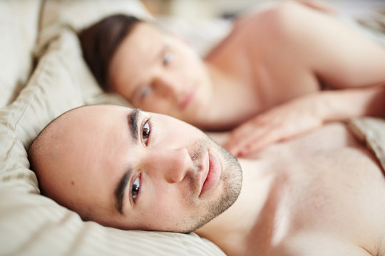 Bald guy lying in bed with his head on pillow and looking at camera with his partner on background