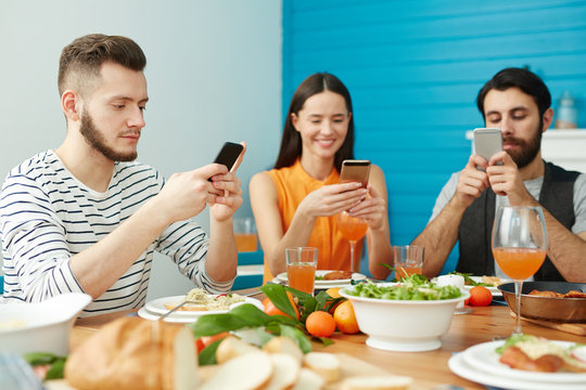 Three young people with gadgets searching in web while sitting by festive table after dinner