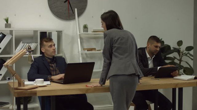 Beautiful brunette businesswoman flirting near the table at workplace in office, man sitting at the desk and looking at sexy standing colleague. Relationship at work and sexual harassment concept.