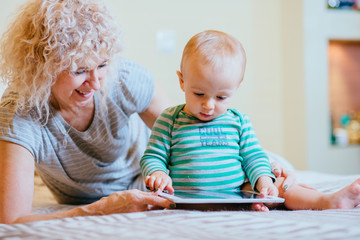 Little cute infant baby boy with curly blond female nanny working with tablet while siting in bedroom at cozy home interior. Real people life and different generations concept.