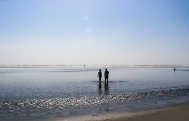 Couple silhouette wading in the water near sunset at the beach with other people walking farther out- sun sparkling off water